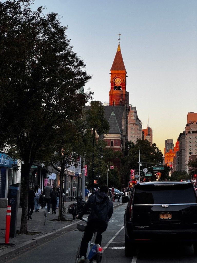 red clock tower with the orange light of the sunset on it, below it trees and cars and a bike lane where a biker is merging into. On the sidewalk, a Citibank and a few other shops with a relatively crowded walkway, with about 7 people clustered together.