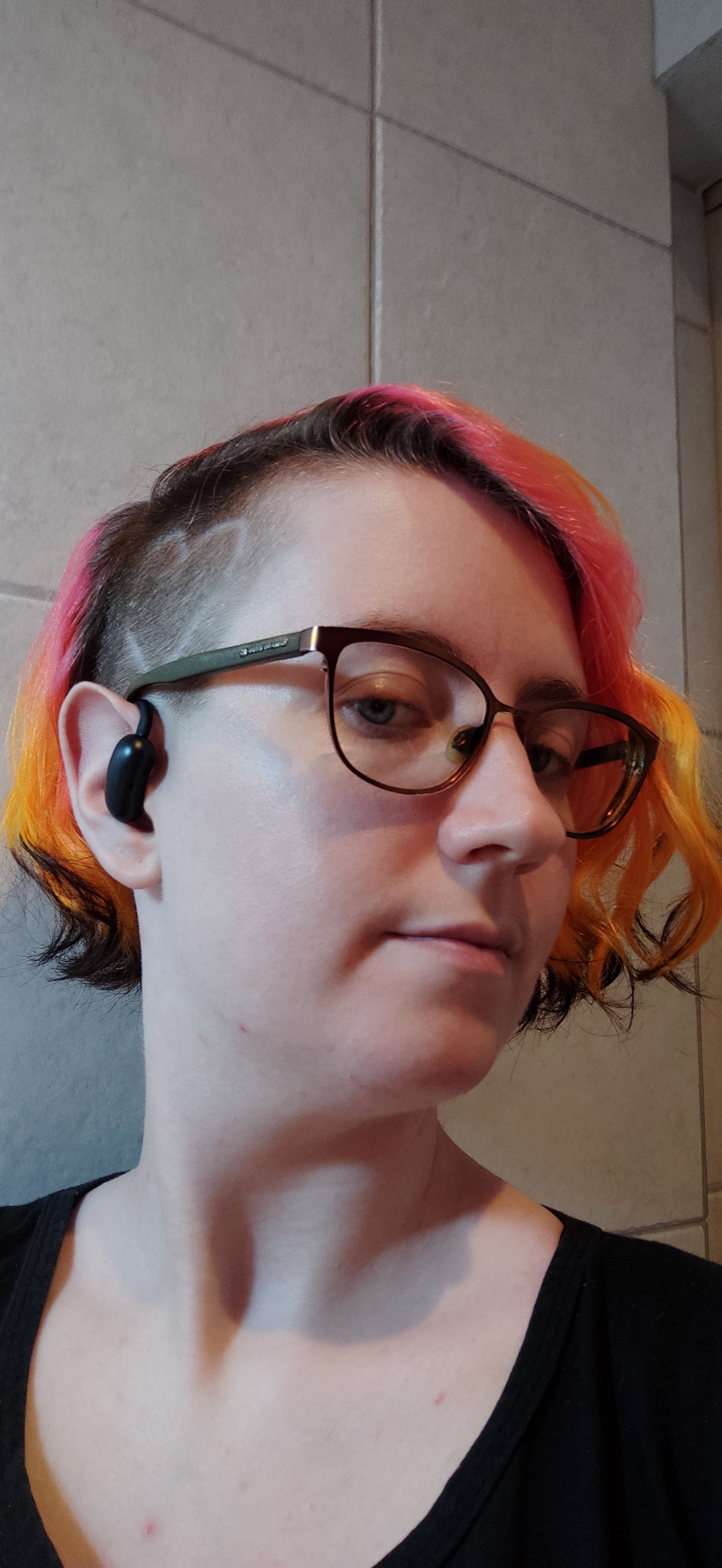 Selfie mostrando meu sidecut recém raspado com um coração desenhado com a máquina. Pessoa branca, olhos azuis, óculos grandes, olhando para a direita, cabelo acima do queixo ondulado, todo jogado para o lado oposto da câmera, vai do rosa para laranja neon, com pontinhas pretas, minha raíz está sem retocar na cor de loiro escuro. Estou usando um fone de ouvido sem fio preto.