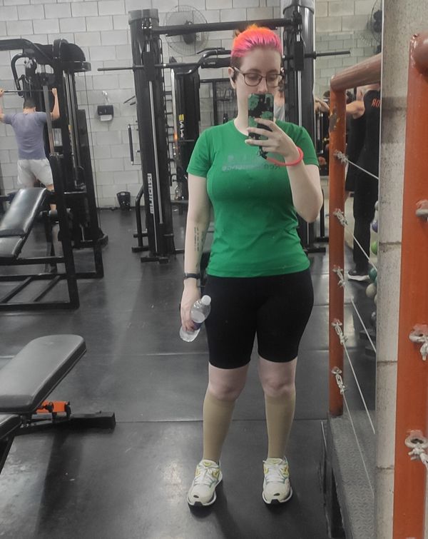 Selfie no espelho da academia, atrás de mim vários equipamentos e duas pessoas malhando. Pessoa branca, usando uma camiseta verde da academia, bermuda colada preta, meias elásticas de compressão na panturrilha na cor bege, tênis esportivo branco com detalhes amarelo, azul e rosa, seguro meu celular com uma capinha preta com dinossauros verde e um cordão laranja neon na mão direita, e uma garrafa de água descartável na mão esquerda, meu cabelo está amarrado para trás só podendo ver a cor rosa e laranja dele, e uso óculos grandes.