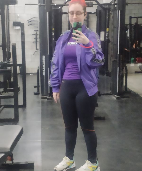 Selfie no espelho da academia, atrás de mim vários equipamentos. Pessoa branca, usando uma camiseta roxa da academia, jaqueta roxa da Twitch aberta, legging preta com linhas de costura laranja, tênis esportivo branco com detalhes amarelo, azul e rosa, seguro meu celular com uma capinha preta com dinossauros verde e um cordão laranja neon, meu cabelo está amarrado para trás só podendo ver a cor rosa e laranja dele, e uso óculos grandes.
